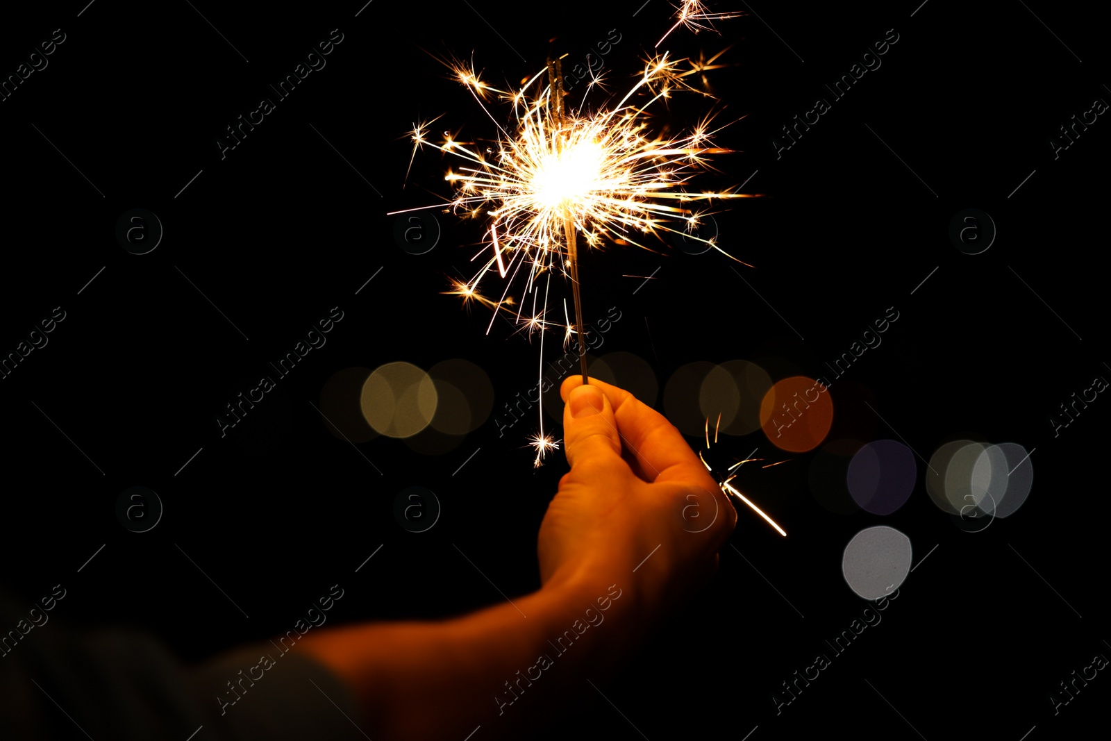 Photo of Woman holding bright burning sparklers against blurred lights, closeup