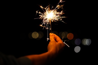 Photo of Woman holding bright burning sparklers against blurred lights, closeup