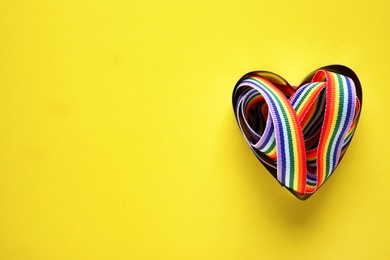 Top view of rainbow ribbon in heart shaped box on yellow background, space for text. Gay symbol