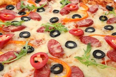 Photo of Tasty pizza with cheese, dry smoked sausages, olives, chili pepper and arugula as background, closeup