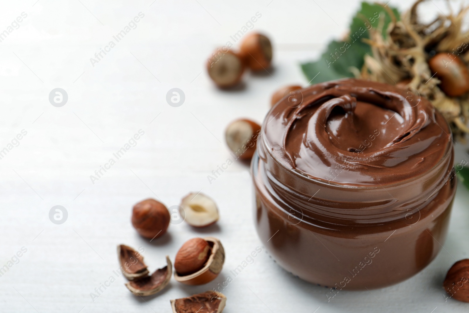 Photo of Glass jar with tasty chocolate spread and hazelnuts on white wooden table, space for text