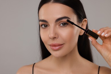 Beautiful young woman applying mascara on grey background, closeup