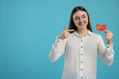 Happy woman pointing at credit card on light blue background, space for text