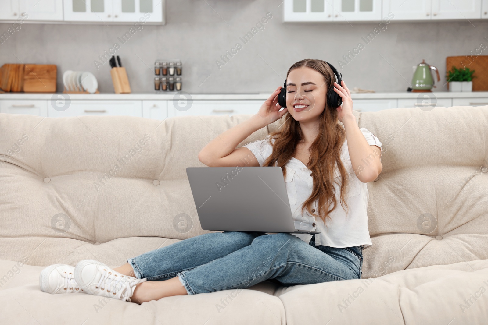 Photo of Happy woman with laptop listening to music in headphones on couch indoors