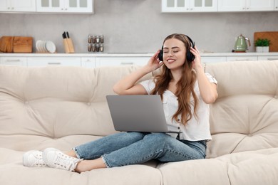 Happy woman with laptop listening to music in headphones on couch indoors