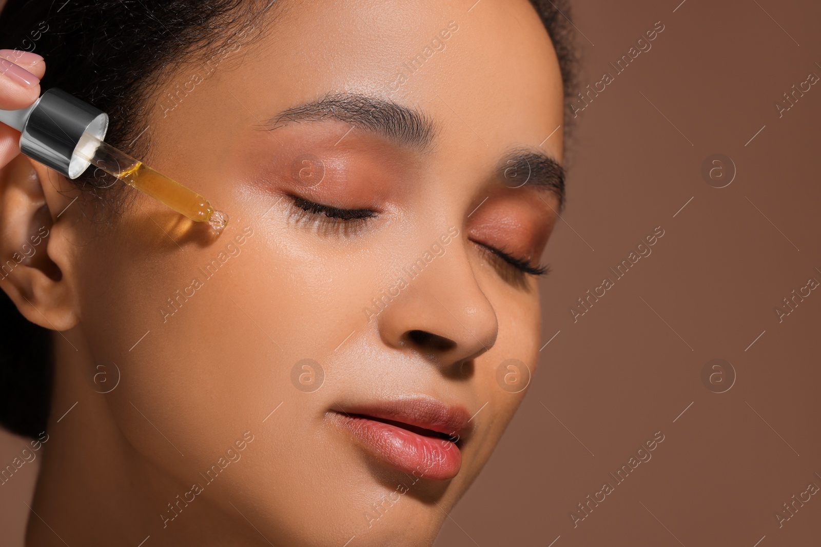 Photo of Beautiful woman applying serum onto her face on brown background, closeup. Space for text