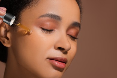 Beautiful woman applying serum onto her face on brown background, closeup. Space for text