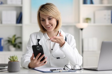 Photo of Smiling doctor with smartphone having online consultation at table in office