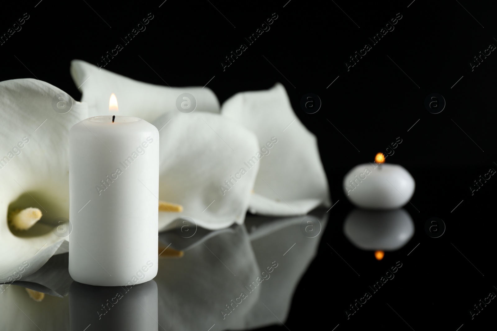 Photo of White calla lily flowers and burning candles on black mirror surface in darkness, closeup with space for text. Funeral symbols