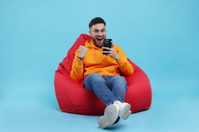 Photo of Happy young man using smartphone on bean bag chair against light blue background
