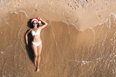 Image of Young woman wearing Santa hat and bikini on sunny beach, top view. Christmas vacation
