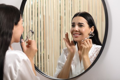 Photo of Emotional young woman with eyelash curler near mirror indoors