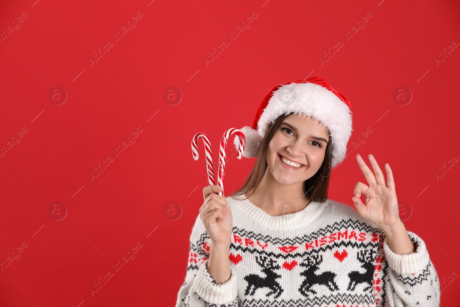 Photo of Pretty woman in Santa hat and Christmas sweater holding candy canes on red background, space for text