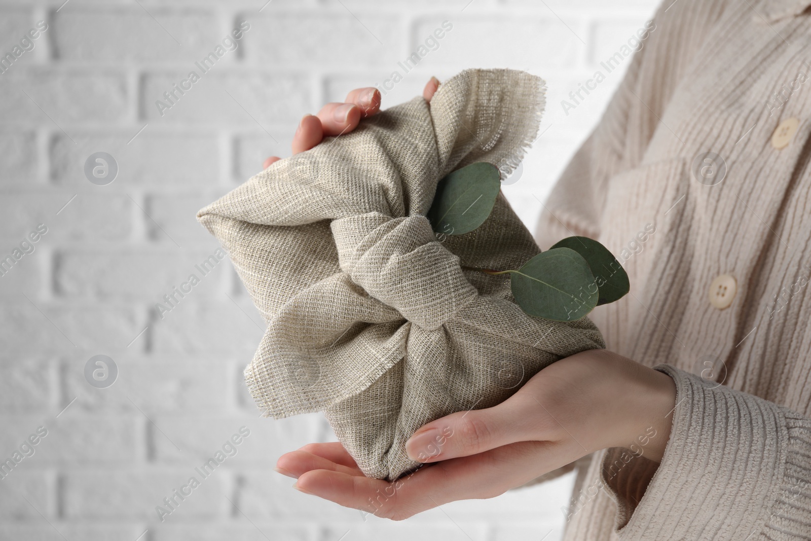 Photo of Furoshiki technique. Woman holding gift packed in fabric and decorated with eucalyptus branch against white wall, closeup