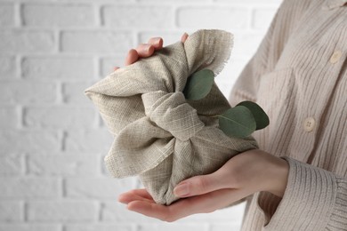 Furoshiki technique. Woman holding gift packed in fabric and decorated with eucalyptus branch against white wall, closeup