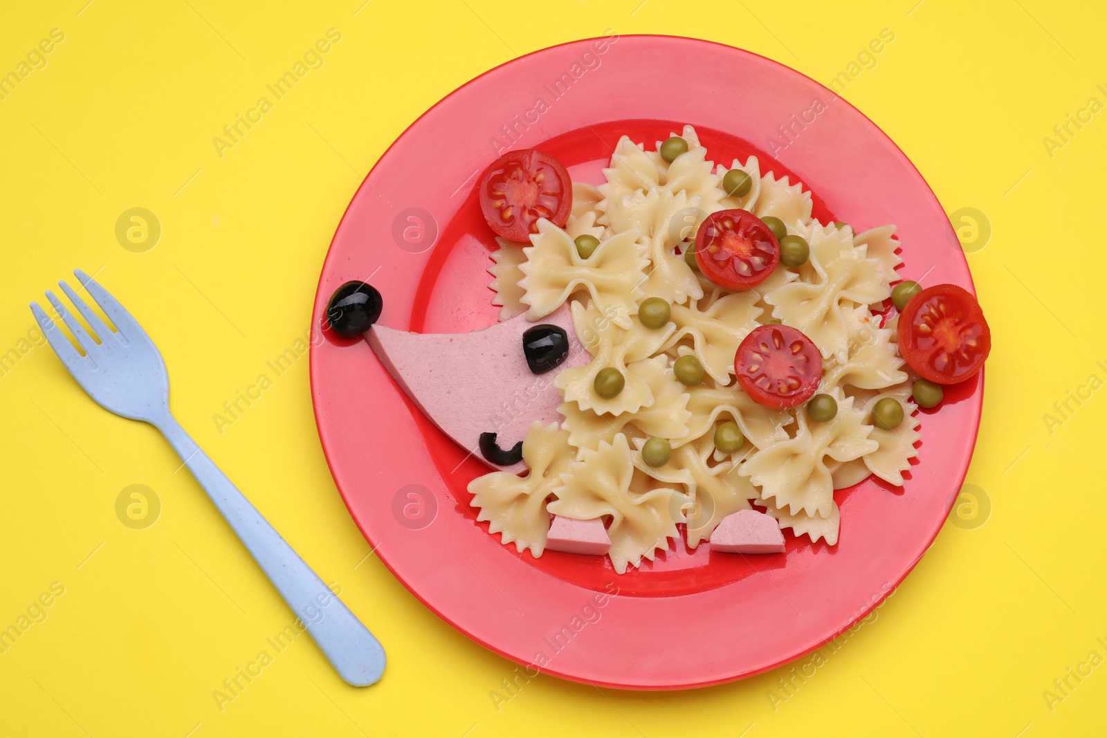 Photo of Creative serving for kids. Plate with cute hedgehog made of delicious pasta, sausages and tomatoes on yellow table, flat lay