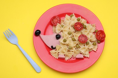 Photo of Creative serving for kids. Plate with cute hedgehog made of delicious pasta, sausages and tomatoes on yellow table, flat lay