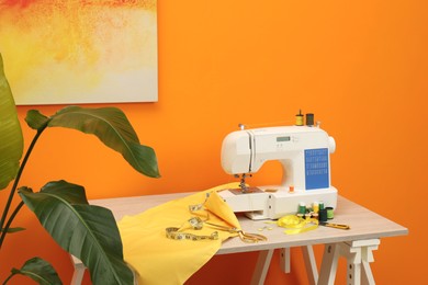 Photo of Modern sewing machine with cloth and craft accessories on wooden table near orange wall in room