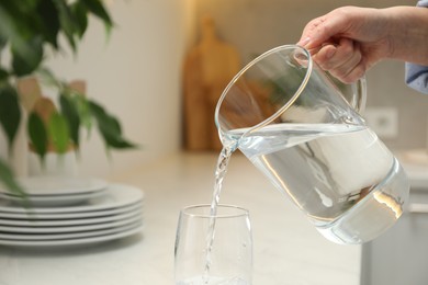 Photo of Woman pouring water from jug into glass at white table in kitchen, closeup