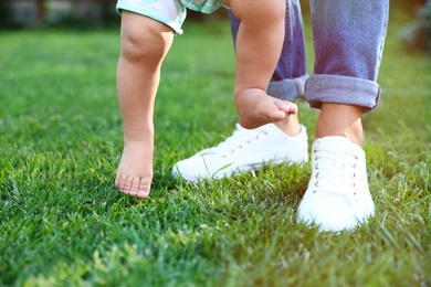 Photo of Cute little baby learning to walk with his nanny on green grass outdoors, closeup