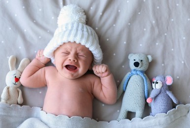 Cute newborn baby wearing white knitted hat with toys in bed, top view