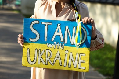 Photo of Young woman holding poster in colors of national flag and words Stand with Ukraine outdoors, closeup