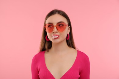 Pink look. Beautiful woman in heart shaped sunglasses blowing bubble gum on color background