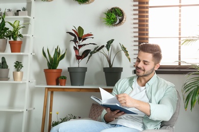 Photo of Young man reading book in room with different home plants