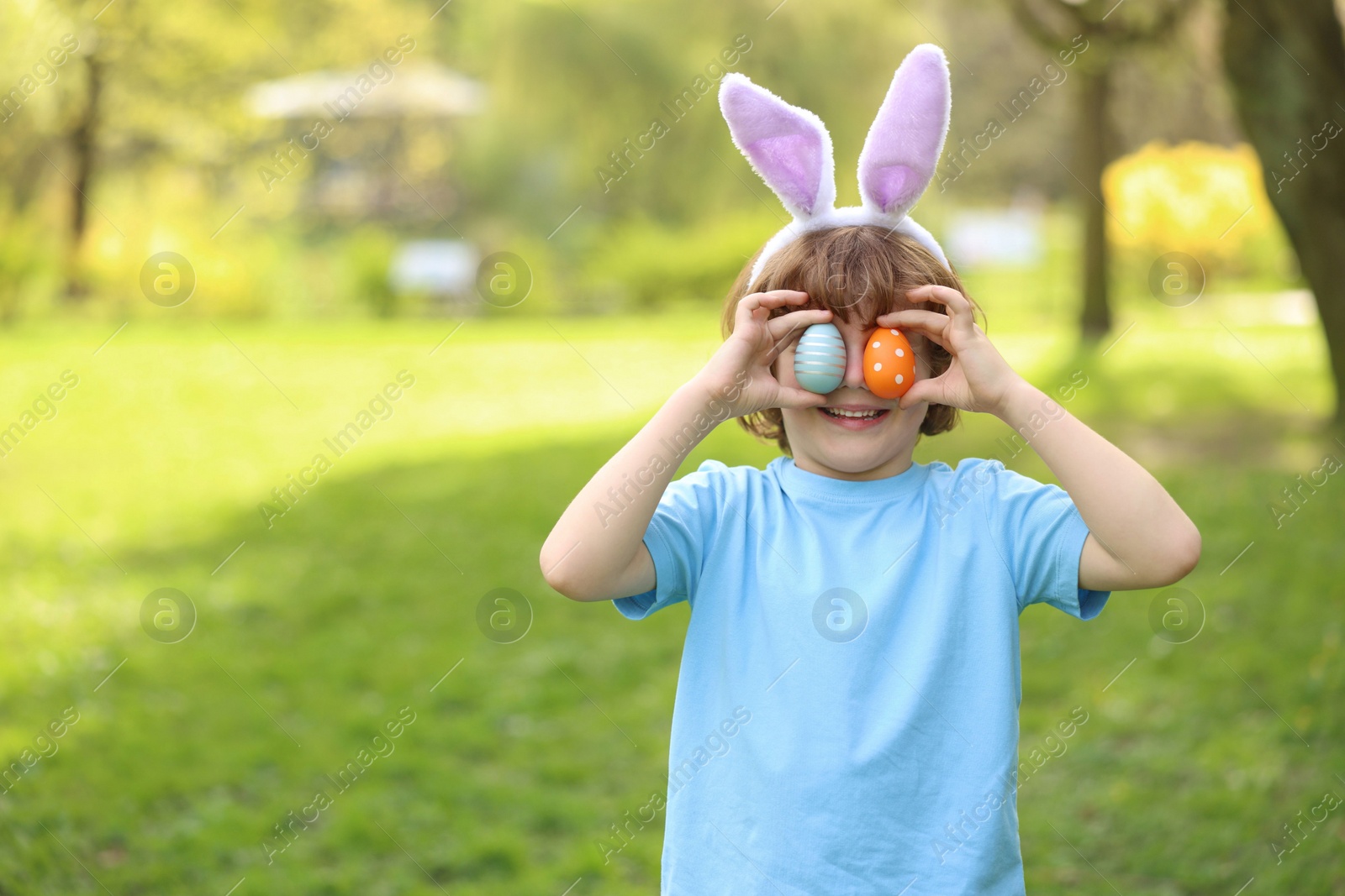 Photo of Easter celebration. Cute little boy in bunny ears covering eyes with painted eggs outdoors, space for text