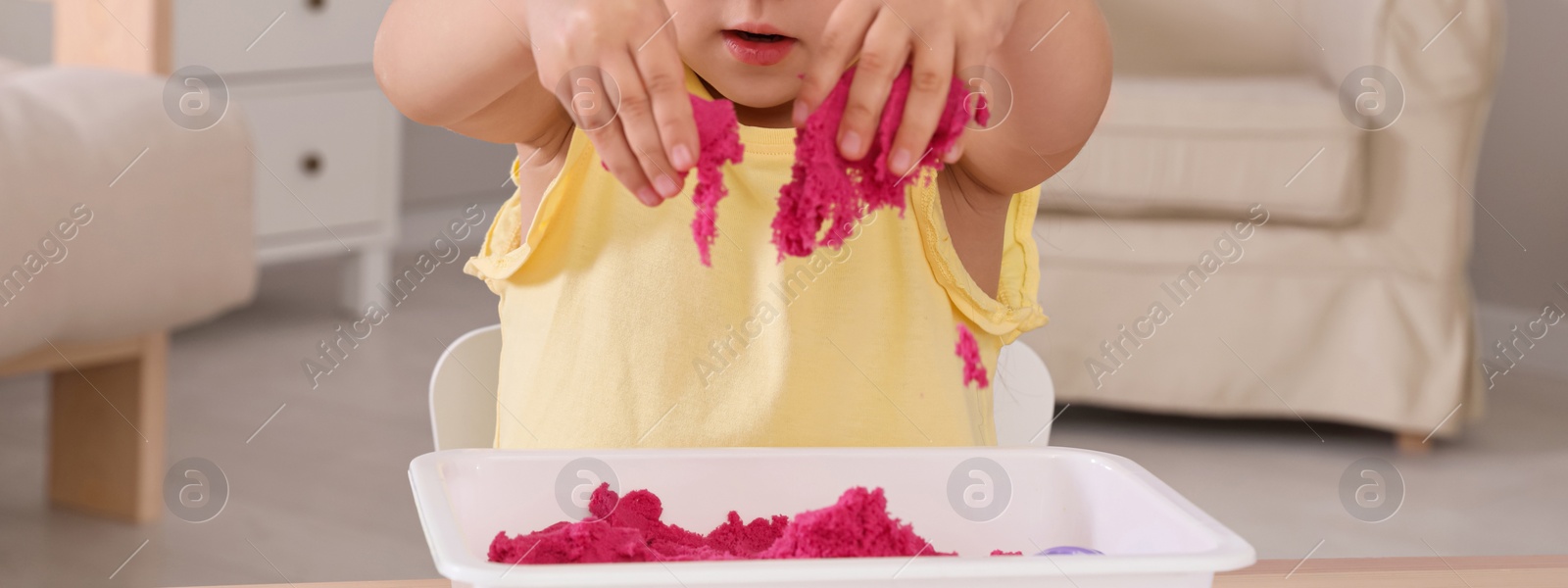 Image of Cute little girl playing with bright kinetic sand at table in room, closeup. Banner design