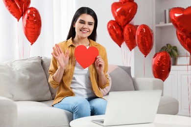 Valentine's day celebration in long distance relationship. Woman holding red paper heart while having video chat with her boyfriend via laptop at home