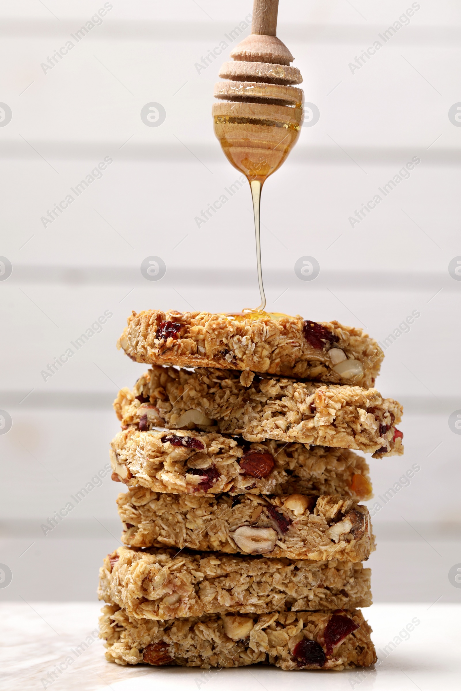 Photo of Pouring honey from dipper onto stack of tasty granola bars on white table