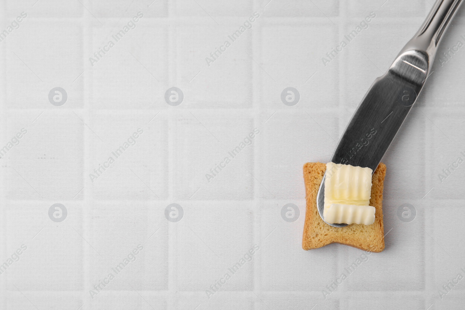 Photo of Tasty butter curl, knife and piece of dry bread on white tiled table, top view. Space for text