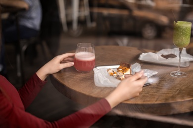 Young woman with cocktail and cake at table in cafe, view from outdoors through window