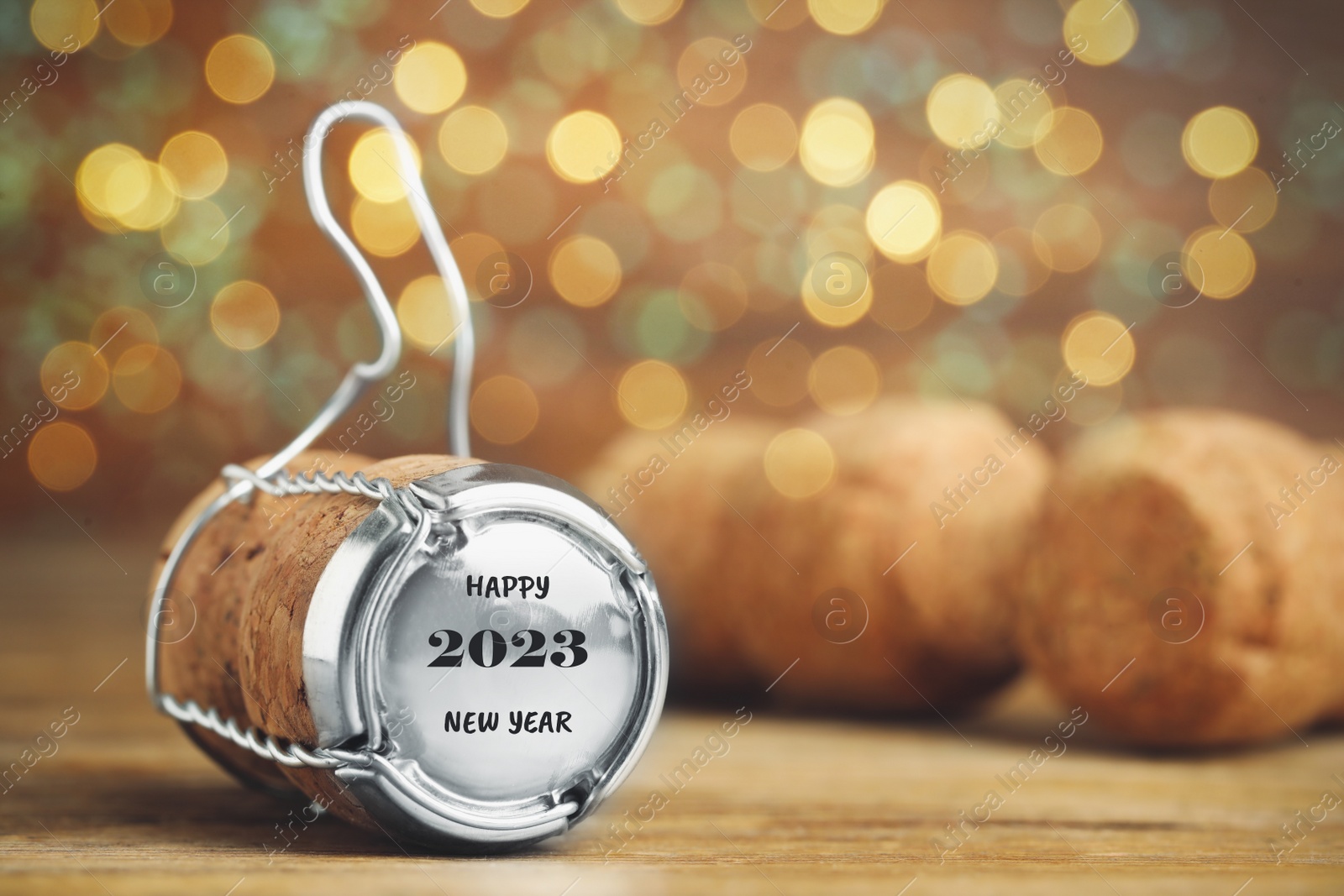 Image of Cork of sparkling wine and muselet cap with engraving Happy 2023 New Year on wooden table against blurred festive lights, space for text. Bokeh effect