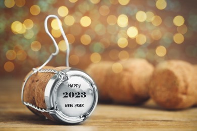 Cork of sparkling wine and muselet cap with engraving Happy 2023 New Year on wooden table against blurred festive lights, space for text. Bokeh effect