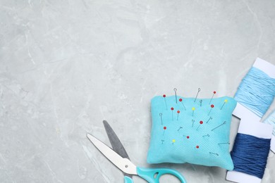 Light blue pincushion with sewing pins, scissors and threads on grey table, flat lay. Space for text