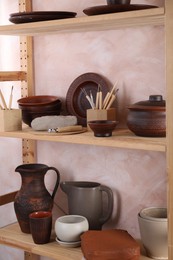 Photo of Set of different crafting tools and clay dishes on wooden rack in workshop