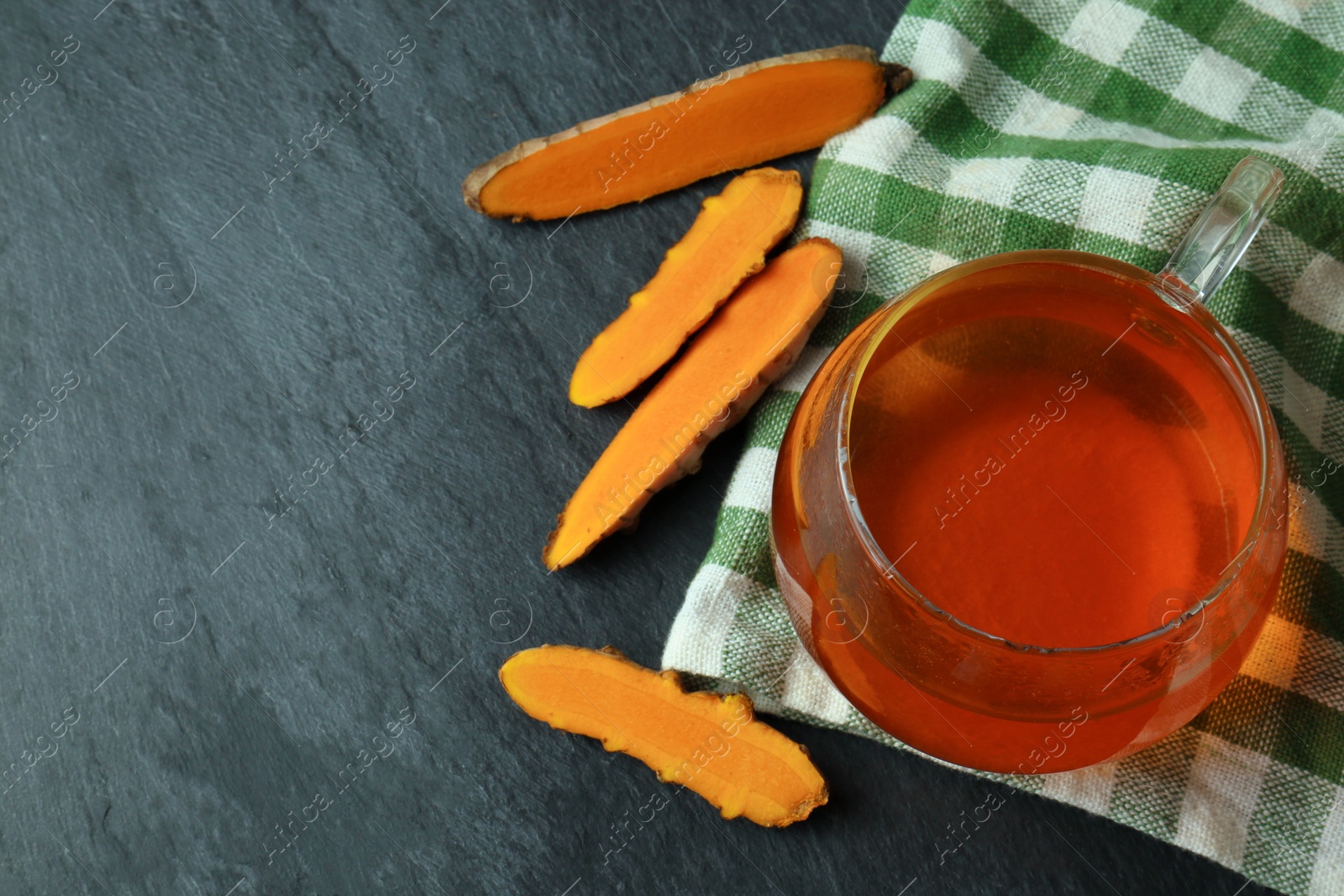 Photo of Glass cup of tasty tea and cut turmeric roots on black textured table, flat lay. Space for text