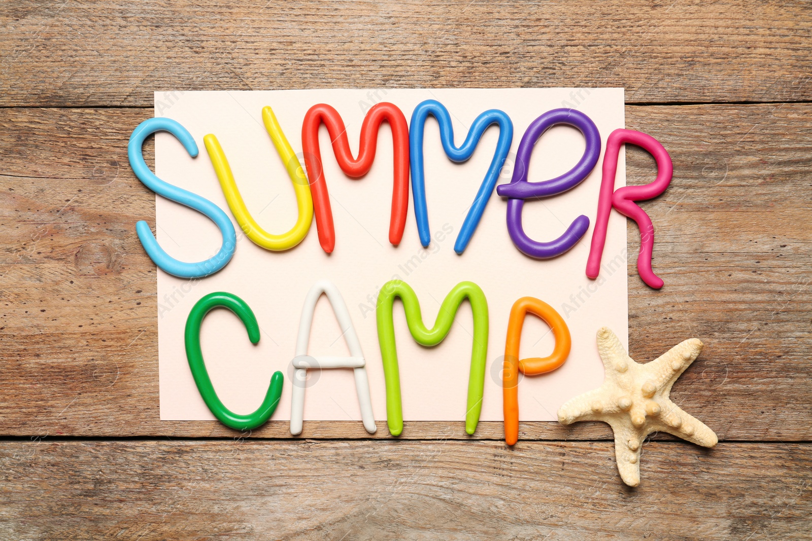 Photo of Text SUMMER CAMP made of modelling clay and starfish on wooden table, flat lay