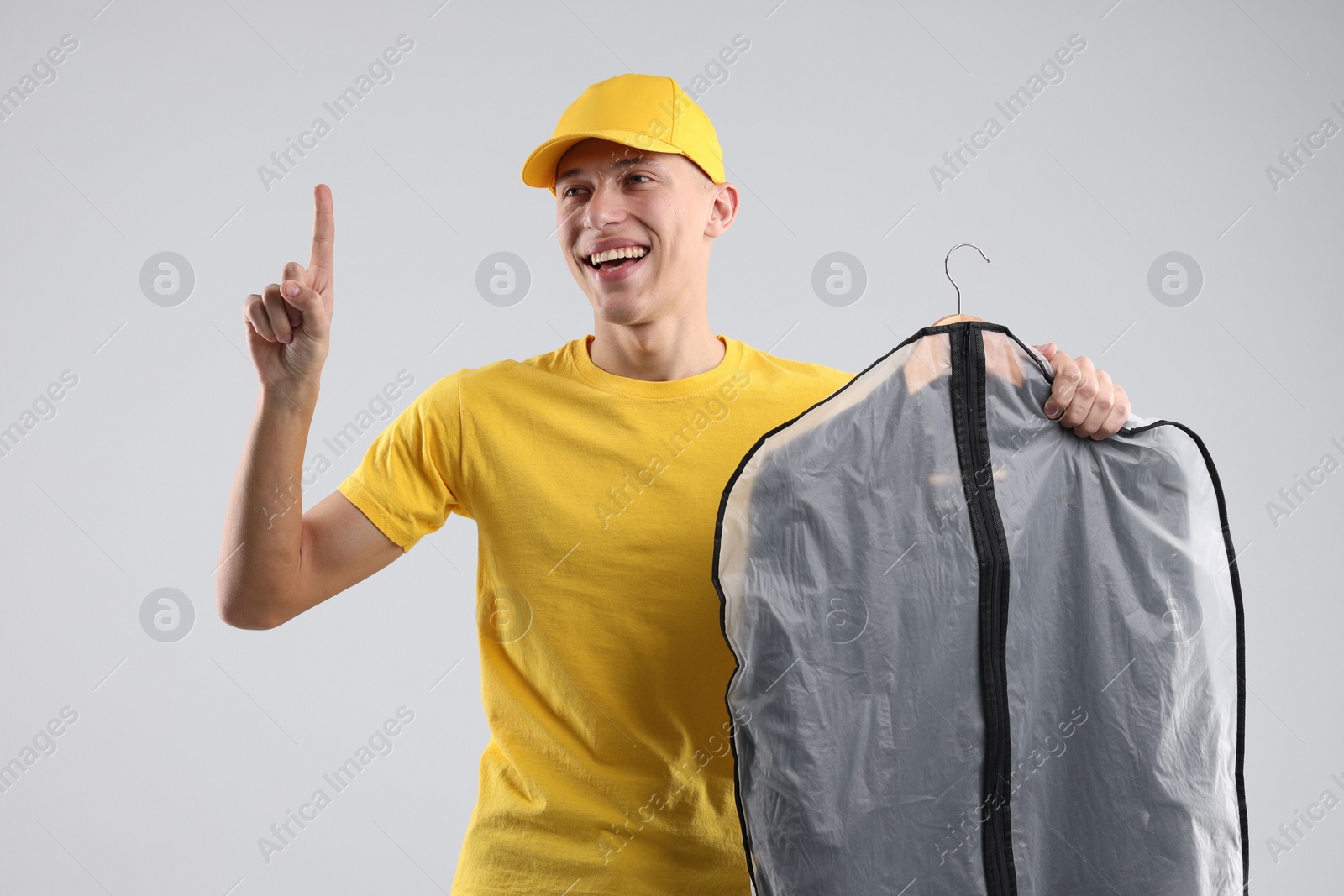Photo of Dry-cleaning delivery. Happy courier holding garment cover with clothes and pointing at something on light grey background
