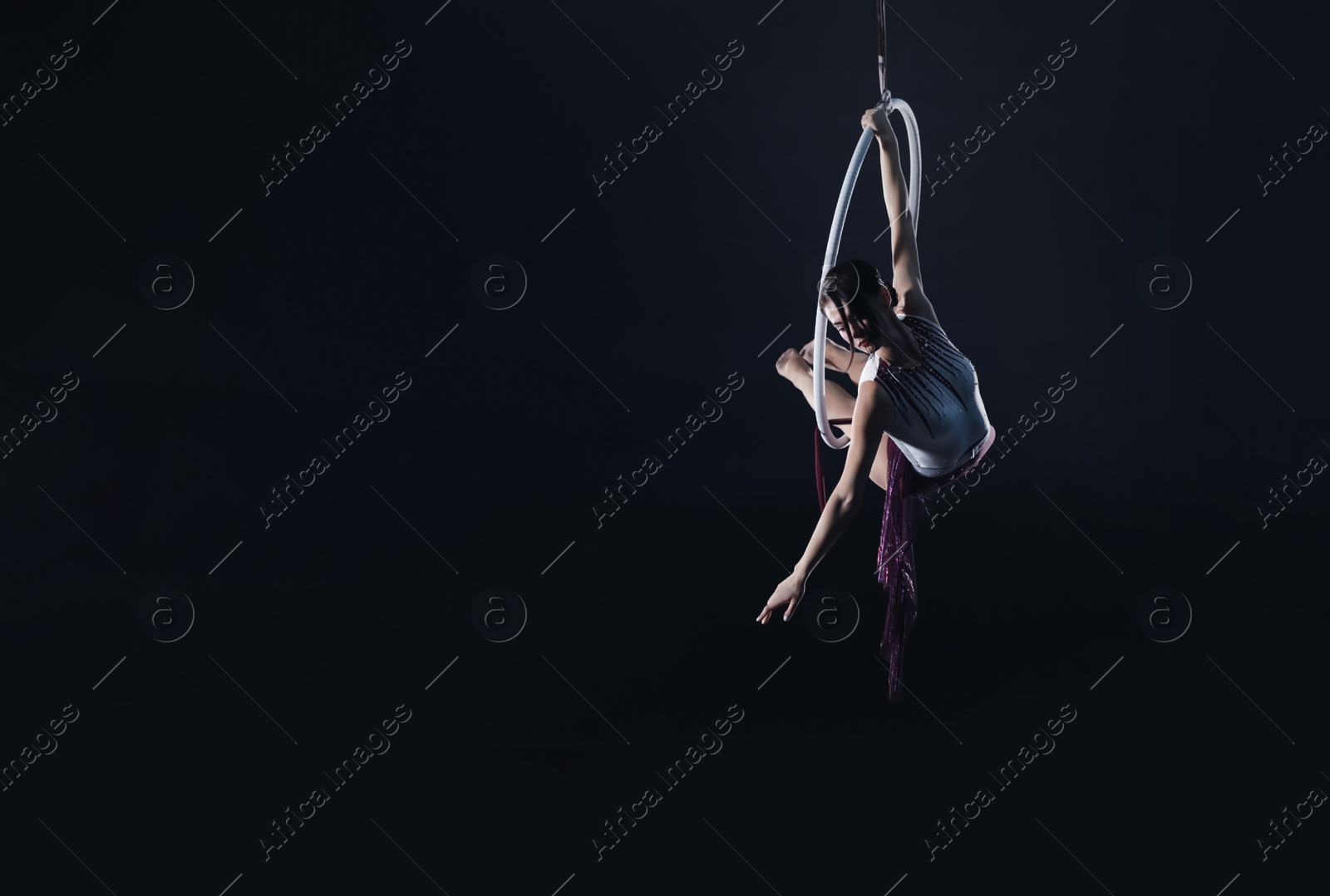 Photo of Young woman performing acrobatic element on aerial ring against dark background. Space for text