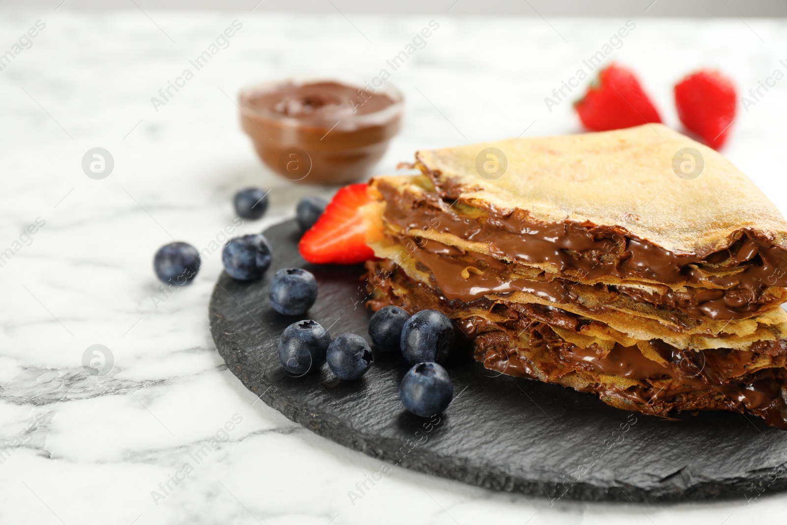 Photo of Delicious thin pancakes with chocolate paste and blueberries on white marble table, closeup