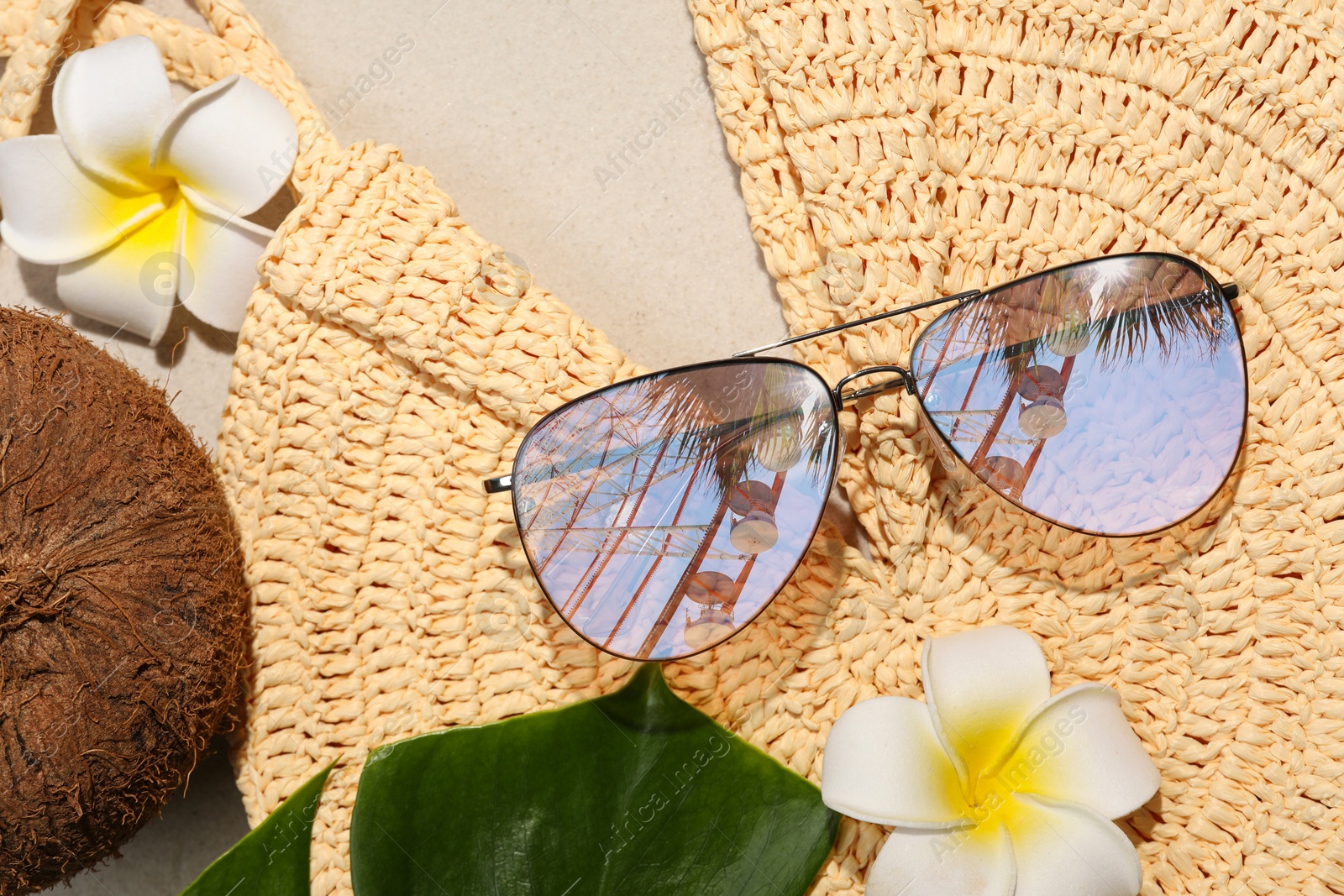 Image of Flat lay composition with stylish sunglasses and wicker bag on sand. Sky, palm tree and observation wheel reflecting in lenses