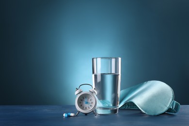 Photo of Alarm clock, soporific pills, sleeping mask and glass of water on blue wooden table, space for text. Insomnia treatment