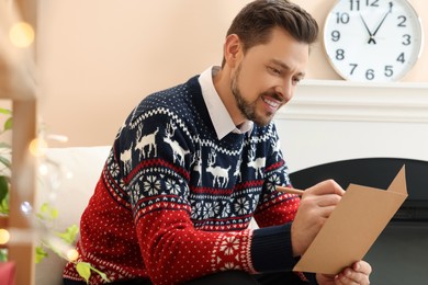 Photo of Happy man writing wishes in Christmas greeting card in living room