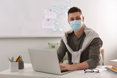 Teacher with protective mask and laptop sitting at desk in classroom. Reopening after Covid-19 quarantine