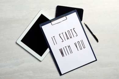Photo of Clipboard with phrase It Starts With You, tablet and smartphone on white table , flat lay