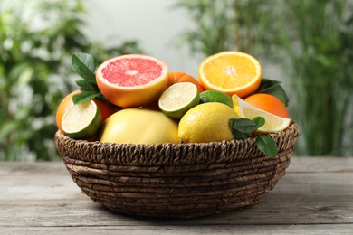 Photo of Different fresh citrus fruits and leaves in wicker basket on wooden table against blurred background, closeup