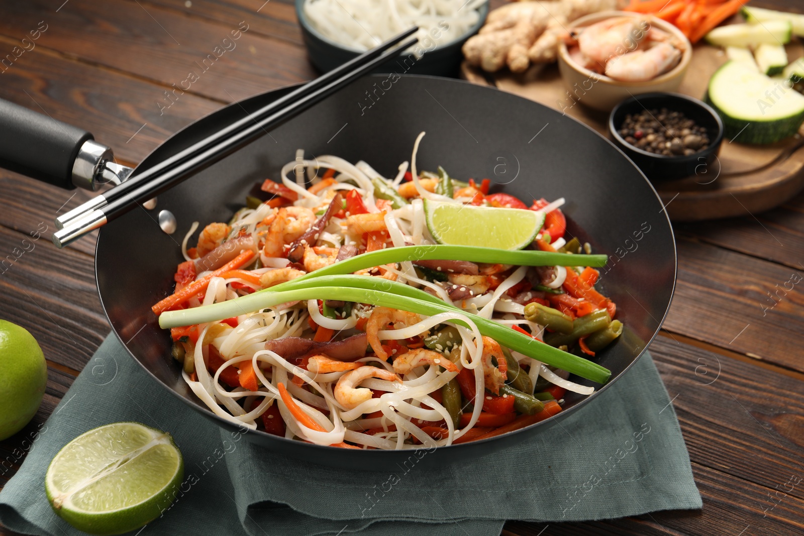 Photo of Shrimp stir fry with noodles and vegetables in wok on wooden table